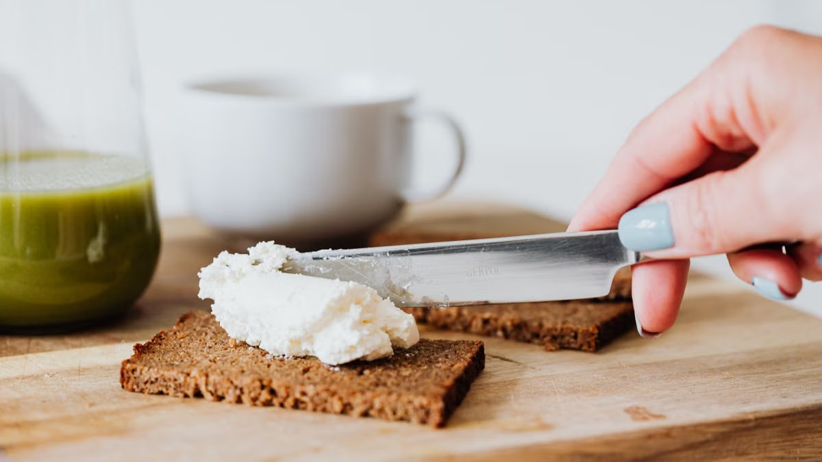 Hand spreading cream cheese on rye bread with a green juice and coffee nearby on a wooden chopping board.